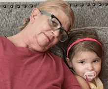 a little girl with a pacifier in her mouth sits next to an older woman wearing glasses