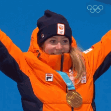 a woman wearing an orange jacket and a black beanie is holding up her arms and wearing a gold medal around her neck