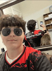 a man wearing sunglasses and a frantik flow shirt takes a picture of himself