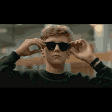 a young man wearing sunglasses and a black sweater is putting on his sunglasses .