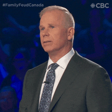 a man in a suit and tie stands in front of a blue background with the words family feud canada above him