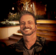 a man with a beard and a camo shirt smiles for the camera