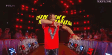 a wrestler in a red shirt is kneeling down in front of a large sign that says shinsuke nakamura .