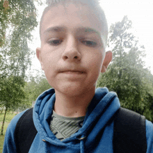 a young boy wearing a blue hoodie and a backpack looks at the camera with trees in the background