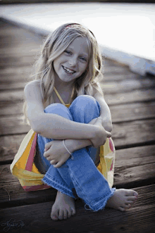 a young girl is sitting on a wooden dock with her legs crossed and smiling