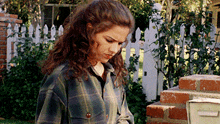 a woman in a plaid shirt is standing in front of a white picket fence next to a mailbox .