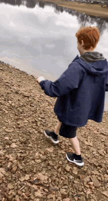 a boy in a blue jacket and nike shoes is walking on a rocky shoreline near a body of water