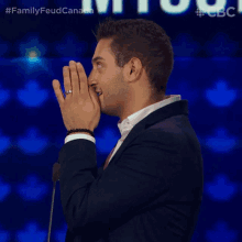 a man in a suit covering his mouth with his hands in front of a microphone with the words family feud canada in the background