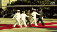 a group of men in military uniforms are walking on a red carpet with the number 32 gun behind them