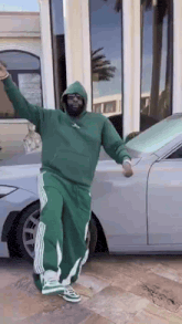 a man in a green hoodie and green pants is standing in front of a silver car .