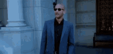 a bald man in a suit and sunglasses is walking in front of a building .