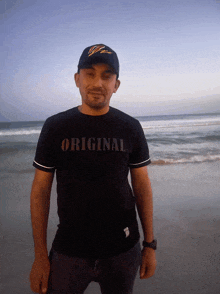 a man wearing a black shirt that says original stands on a beach