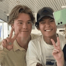 two young men are standing next to each other and giving a peace sign while holding a cell phone .