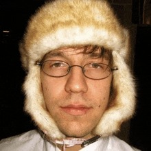 a young man wearing glasses and a fur hat looks at the camera