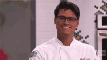 a man wearing glasses and a chef 's jacket is smiling in a kitchen .