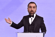 a man in a tuxedo and bow tie stands at a podium giving a speech