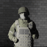 a soldier wearing a helmet and mask holds a bullet proof vest in front of a brick wall