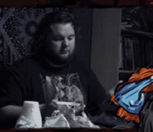 a man with a beard is sitting at a table with a drawing of a man in a black shirt behind him