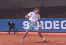 a man in a white shirt and blue shorts is playing tennis in front of a sign that says sardeg