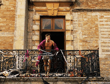 a shirtless man is standing on a balcony with a wrought iron railing and a brick building in the background