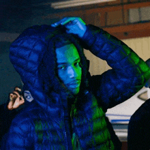 a man wearing a blue jacket with a hood is standing in a dark room with a green light behind him .