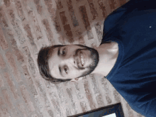 a man with a beard and a blue shirt smiles in front of a brick wall