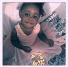 a little girl in a pink dress is sitting on a bed holding a container of popcorn