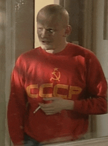 a man wearing a red sweater with a hammer and sickle on it is smoking a cigarette .