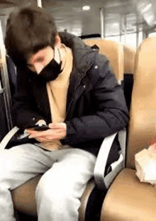 a man wearing a mask is sitting in a chair on a bus looking at his phone .