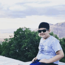 a boy wearing sunglasses and a blue shirt is sitting on a wall .