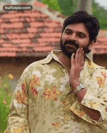 a man with a beard wearing a floral shirt and a watch is standing in front of a tiled roof .