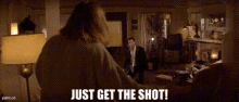 a man standing in a living room with the words `` just get the shot '' written on the bottom .