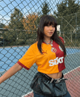 a woman wearing a sixt shirt stands in front of a fence