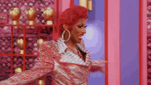 a drag queen with red hair and hoop earrings stands in front of a pink wall