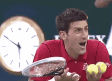 a man in a red shirt is holding a tennis racquet and tennis balls in front of a clock .