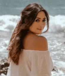 a woman in a white off the shoulder top is standing on the beach looking at the camera .