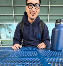 a man wearing glasses and a blue hoodie sitting at a blue metal table