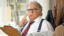 an older man wearing suspenders and a tie is smoking a cigarette while holding a clipboard and a newspaper .