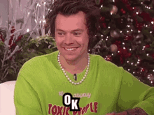 harry styles is wearing a green sweater and a pearl necklace and smiling .