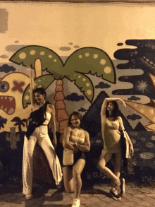 three women pose in front of a wall with a palm tree and a monster on it