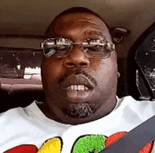 a man wearing glasses and a t-shirt is sitting in a car and making a funny face .