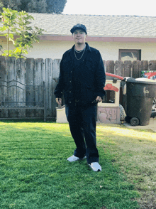 a man in a black jacket and white shoes stands in the grass in front of a fence