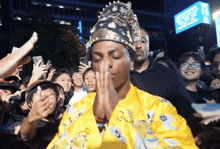 a man wearing a yellow robe and a crown is praying in front of a crowd .