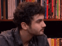 a man with curly hair and a beard is sitting in front of a bookshelf full of books including one called venus