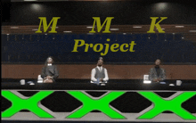 three people are sitting at a table in front of a sign that says mmk project