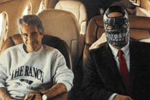 a man wearing a the ranch sweatshirt sits next to a man wearing a mask