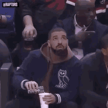 a man with a beard is holding a cup of coffee while sitting in the stands during a basketball game .