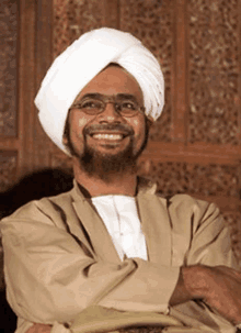 a man wearing a white turban and glasses is smiling and sitting with his arms crossed .