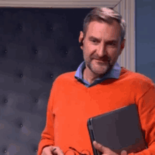 a man in an orange sweater holds a black laptop