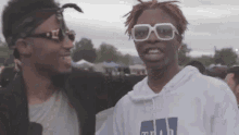 two young men are standing next to each other at a festival . one of the men is wearing sunglasses and a bandana .